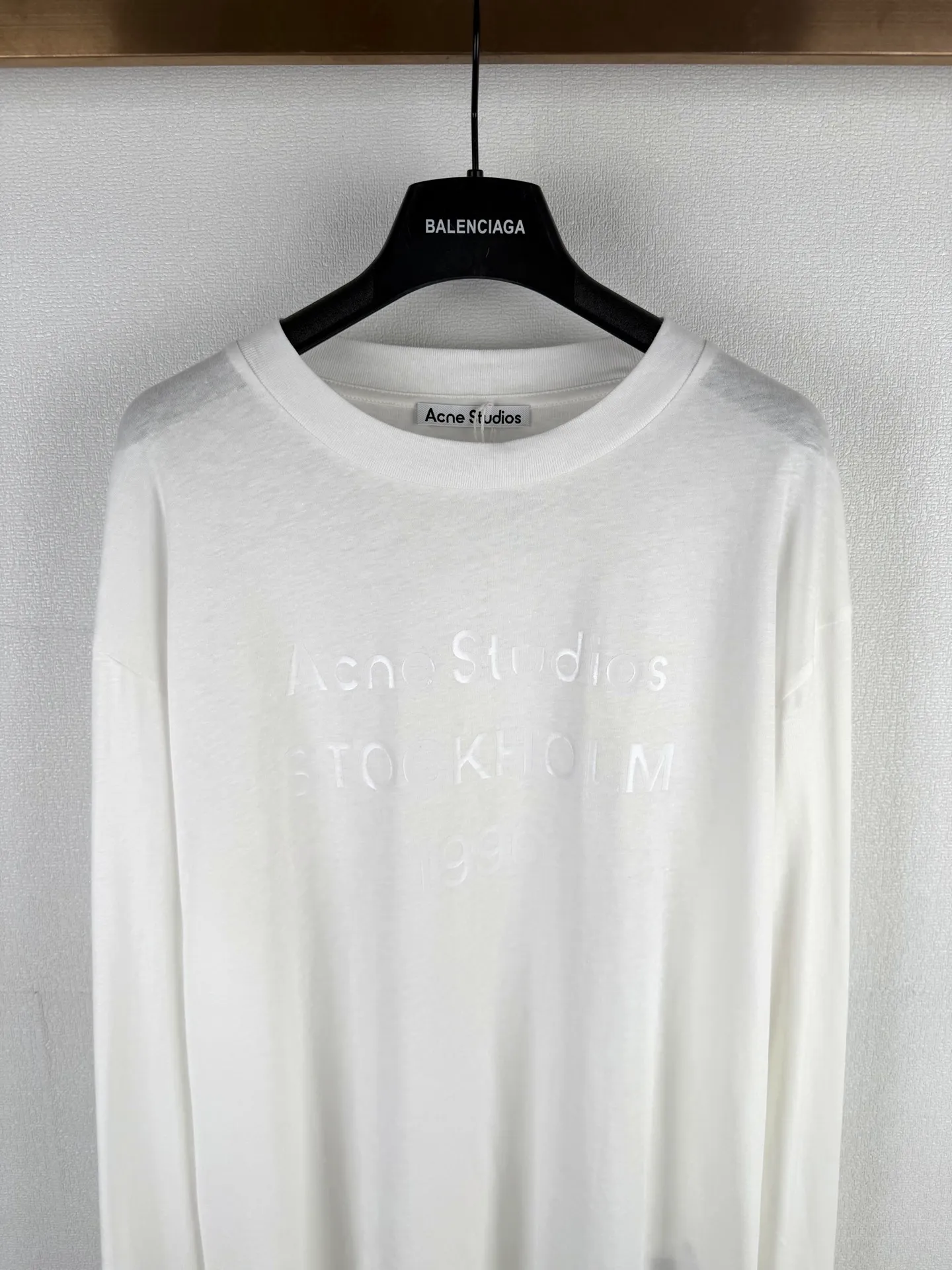 ACNE STUDIOS stamp logo printed long-sleeved T-shirt This long-sleeved round neck style is made of cotton/linen blend jersey fabric with an irregular textured surface for a retro style. Ac stamp logo at front. Relaxed fit, unisex style, hip-length. The fineness of the 32-count yarn ensures a delicate touch, and the introduction of hemp fiber is the finishing touch. It breaks the conventional softness of pure cotton. On the basis of being skin-friendly and soft, it brings natural roughness, knotty particles and micro-wrinkle visual effects, creating an irregular texture of a natural rough base. The fabric is piece-dyed to create a soft, matte finish. Hemp fiber significantly improves moisture wicking properties and provides a dry touch. It has an extremely light and breathable wearing experience. The fabric can naturally drape according to the body contour, forming soft folds, which perfectly serves the casual and loose version, achieving a harmonious unity of material and silhouette. The logo printing on the front is not a simple offset printing, but uses a double-layer process of environmentally friendly elastic glue and varnish. The bottom layer is environmentally friendly elastic glue, which is composed of water-based environmentally friendly resin. After being scraped through screen printing and cured at high temperature, a soft and continuous adhesive film with strong stretch elasticity is formed. This layer of film can perfectly stretch and retract with the fabric, completely solving the problems of cracking and stiffness in traditional printing, and ensuring that the wear on light and thin fabrics does not feel tight. A transparent varnish that is accurately overprinted on the surface creates a high-gloss gloss effect in specific areas, contrasting with the matte or semi-matte texture of the glue. This layer of varnish not only significantly improves the color contrast and three-dimensional layering of the pattern, but also strengthens the wear-resistant and wash-resistant properties, making the printing on the rough hemp base appear elastic, durable and glossy.